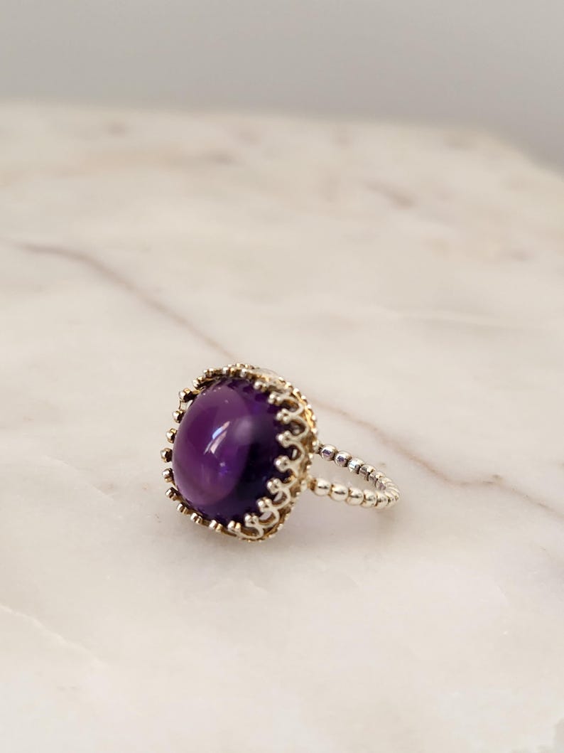 May include: A silver ring with a large, oval, deep purple gemstone. The gemstone is set in a gold-colored, ornate bezel. The ring band is silver and features a beaded design. The ring is displayed on a white marble surface.