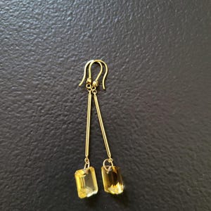 May include: A pair of gold-toned drop earrings featuring long, slender metal bars suspended from curved ear wires. Each bar terminates in a rectangular, faceted yellow gemstone that catches the light, creating a clear and reflective appearance.