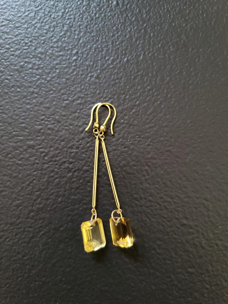 May include: A pair of gold-toned drop earrings featuring long, slender metal bars suspended from curved ear wires. Each bar terminates in a rectangular, faceted yellow gemstone that catches the light, creating a clear and reflective appearance.
