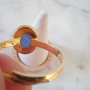 K&ouml;nnte beinhalten: Ein goldener Ring mit einem blauen ovalen Edelstein, der in einer Zargenfassung gefasst ist. Der Ring befindet sich auf einem h&ouml;lzernen Ringst&auml;nder.