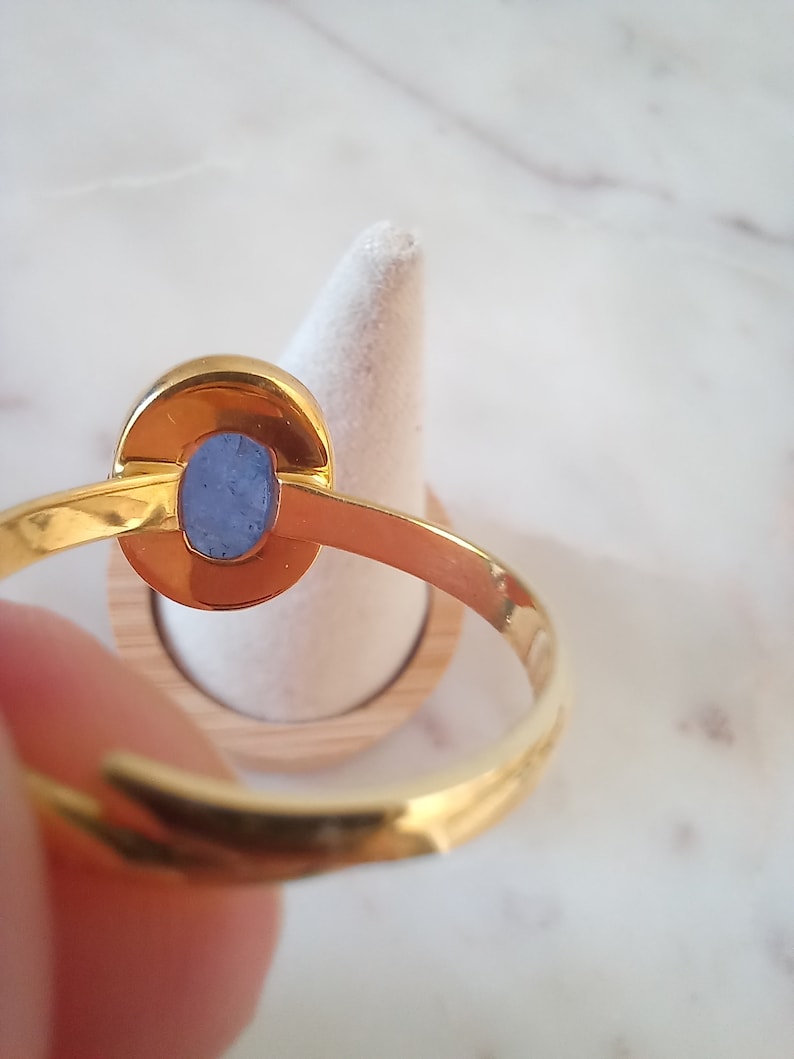 K&ouml;nnte beinhalten: Ein goldener Ring mit einem blauen ovalen Edelstein, der in einer Zargenfassung gefasst ist. Der Ring befindet sich auf einem h&ouml;lzernen Ringst&auml;nder.