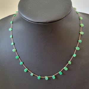 May include: A delicate necklace featuring small, teardrop-shaped green gemstones strung on a thin, light-brown cord. The necklace is displayed on a black mannequin, showcasing the jewelry's design and color.