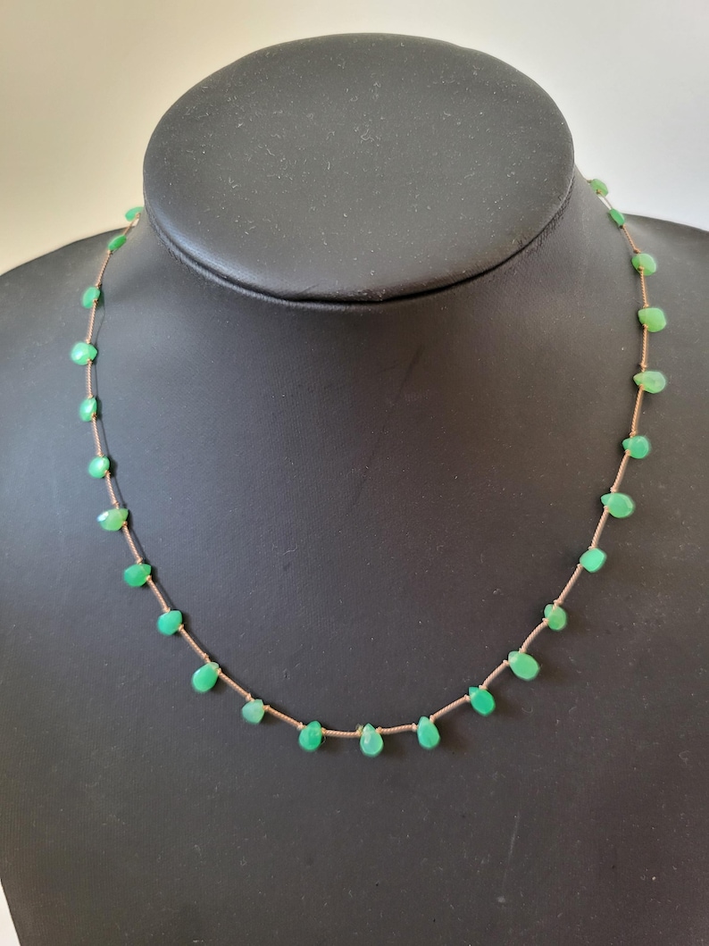 May include: A delicate necklace featuring small, teardrop-shaped green gemstones strung on a thin, light-brown cord. The necklace is displayed on a black mannequin, showcasing the jewelry's design and color.