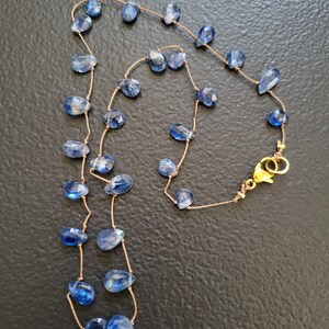 Puede incluir: Un collar delicado que presenta numerosas piedras preciosas de cianita azul peque&ntilde;as, facetadas y con forma de l&aacute;grima, ensartadas en un cord&oacute;n fino de color marr&oacute;n claro. El collar se asegura con un cierre de mosquet&oacute;n de metal dorado.