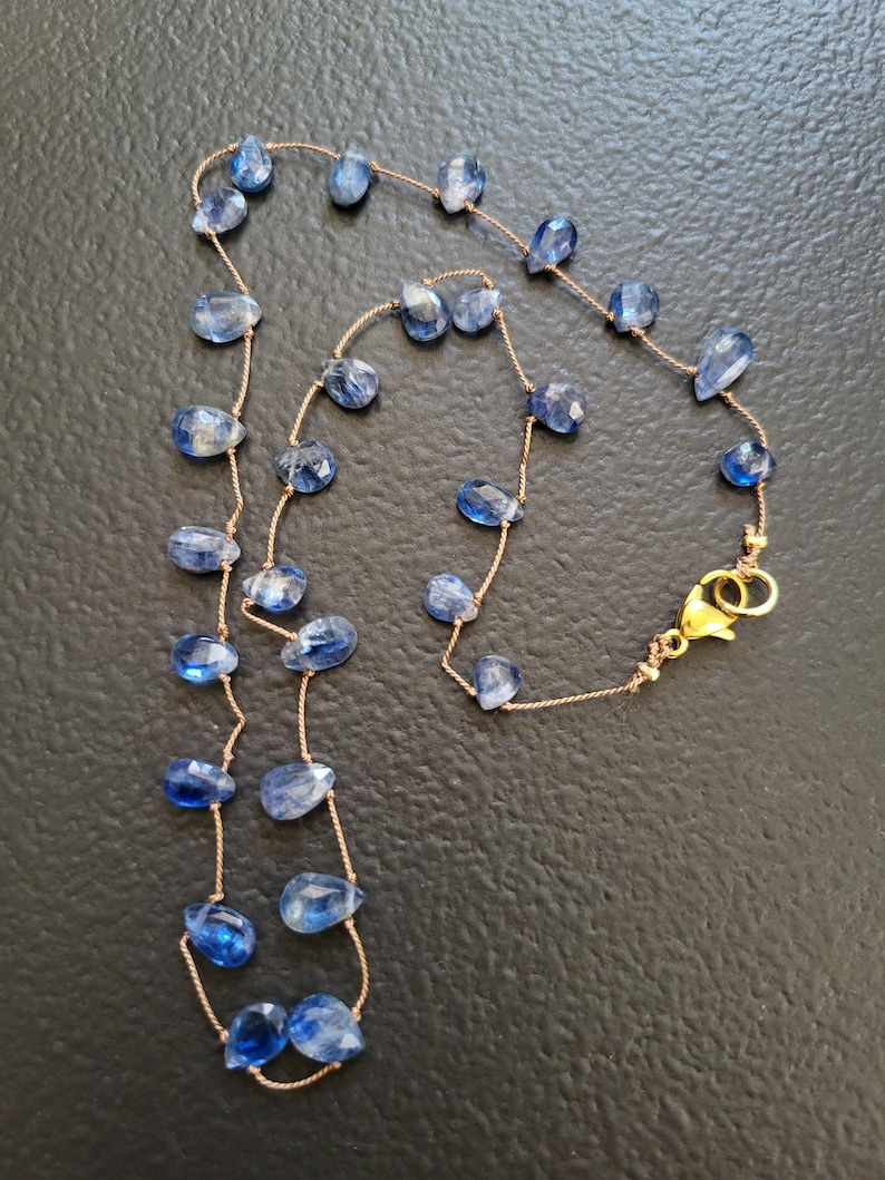 Puede incluir: Un collar delicado que presenta numerosas piedras preciosas de cianita azul peque&ntilde;as, facetadas y con forma de l&aacute;grima, ensartadas en un cord&oacute;n fino de color marr&oacute;n claro. El collar se asegura con un cierre de mosquet&oacute;n de metal dorado.