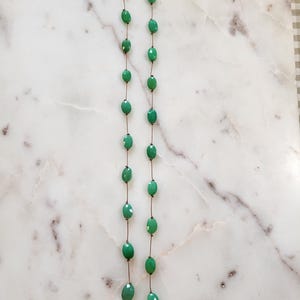 May include: A necklace featuring oval-shaped, faceted green beads strung on a thin, bronze-colored chain. The necklace has a simple design with a clasp at the top. The beads are a vibrant shade of green, and the necklace is displayed on a white and gray marble surface.