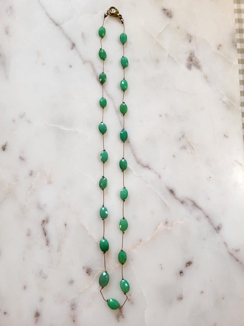 May include: A necklace featuring oval-shaped, faceted green beads strung on a thin, bronze-colored chain. The necklace has a simple design with a clasp at the top. The beads are a vibrant shade of green, and the necklace is displayed on a white and gray marble surface.