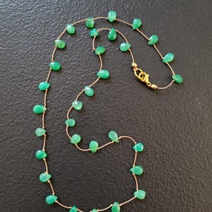 May include: A delicate necklace featuring teardrop-shaped green gemstones strung on a thin, gold-colored chain. The necklace has a gold clasp and is displayed against a dark background. The gemstones are evenly spaced.