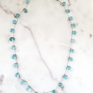 Pu&ograve; includere: Una delicata collana con perline a forma di goccia, di colore azzurro chiaro, infilate su un sottile cordoncino marrone. La collana ha una chiusura color oro ed &egrave; esposta su una superficie di marmo bianco. Le perline sono uniformemente distanziate.