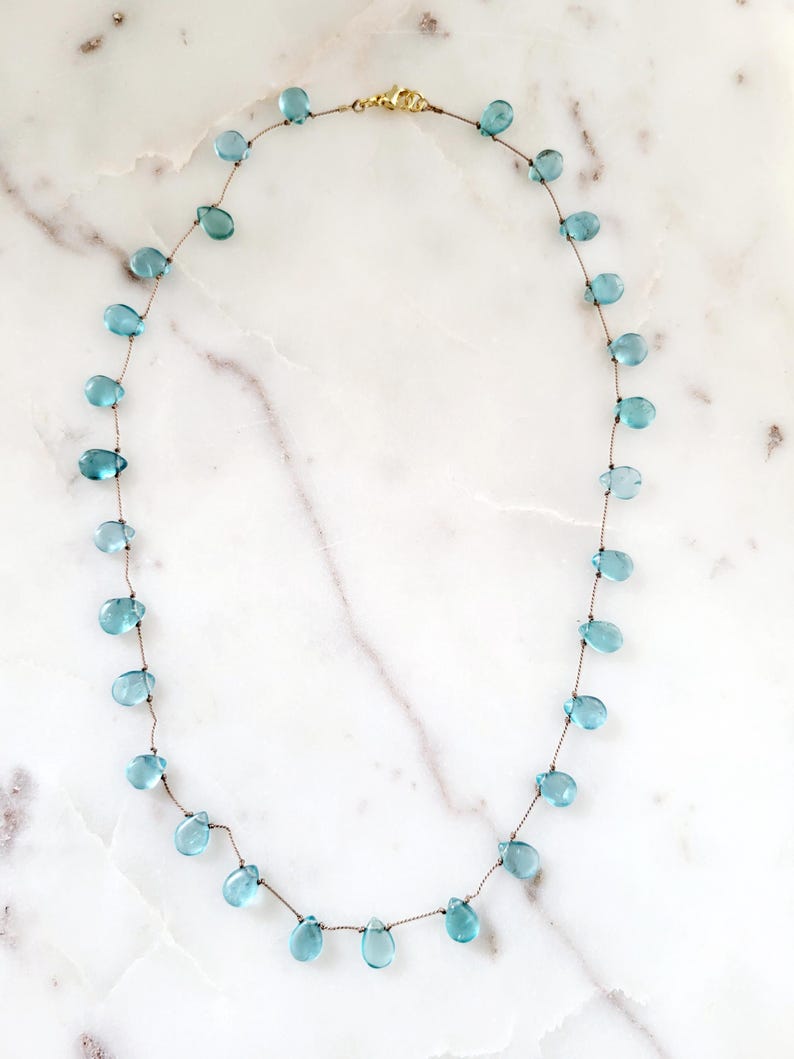 Pu&ograve; includere: Una delicata collana con perline a forma di goccia, di colore azzurro chiaro, infilate su un sottile cordoncino marrone. La collana ha una chiusura color oro ed &egrave; esposta su una superficie di marmo bianco. Le perline sono uniformemente distanziate.