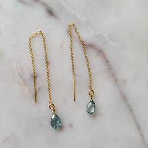 May include: A pair of gold-colored threader earrings with teardrop-shaped, light blue gemstones. The earrings have a delicate chain design with a vertical bar at the top. The gemstones are a soft, pale blue color.
