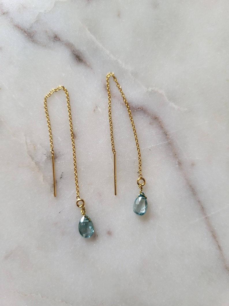 May include: A pair of gold-colored threader earrings with teardrop-shaped, light blue gemstones. The earrings have a delicate chain design with a vertical bar at the top. The gemstones are a soft, pale blue color.