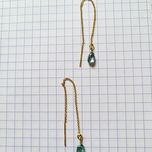 May include: A pair of gold-colored threader earrings. Each earring features a delicate gold chain, a straight bar, and a teardrop-shaped, light blue gemstone. The earrings are displayed on a white grid background.