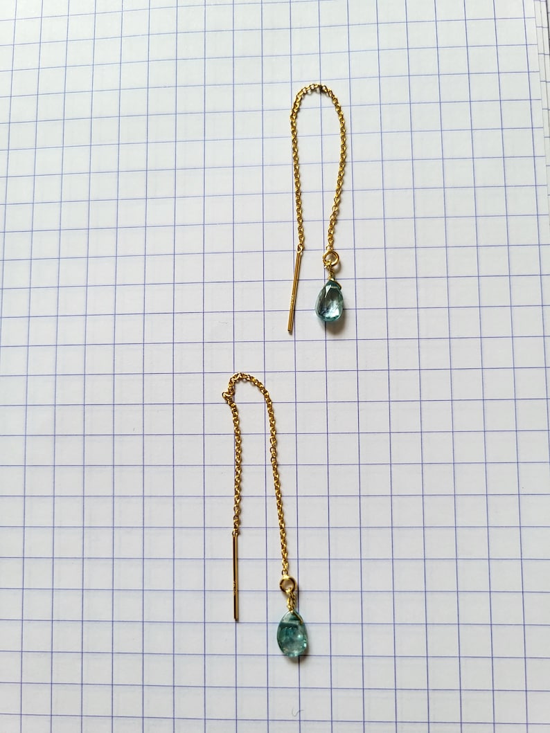 May include: A pair of gold-colored threader earrings. Each earring features a delicate gold chain, a straight bar, and a teardrop-shaped, light blue gemstone. The earrings are displayed on a white grid background.