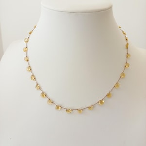 Puede incluir: Un delicado collar con pequeñas piedras preciosas amarillas y redondas ensartadas en un fino cordón marrón. El collar se exhibe en un maniquí blanco, mostrando el diseño y el color de la joya. Las piedras están espaciadas uniformemente.