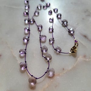 Op de afbeelding: Een delicate ketting met een hanger van paarse edelsteen. De ketting is gemaakt van een dunne paarse koord en heeft een reeks kleine, ronde, paarse edelstenen.