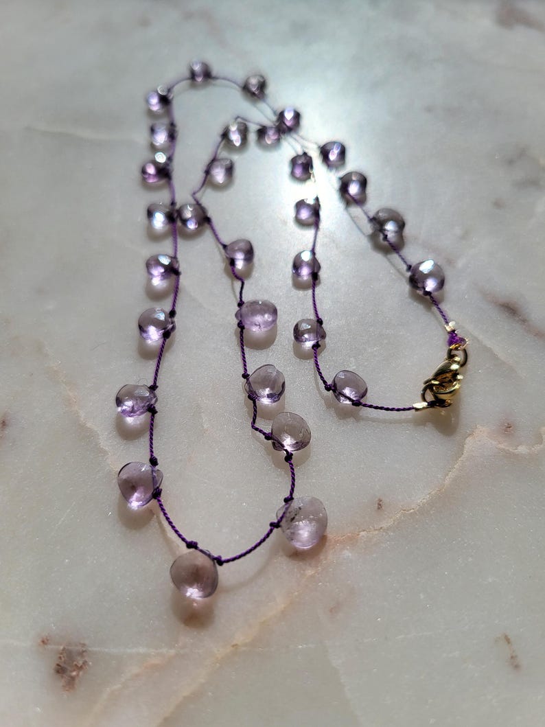 Op de afbeelding: Een delicate ketting met een hanger van paarse edelsteen. De ketting is gemaakt van een dunne paarse koord en heeft een reeks kleine, ronde, paarse edelstenen.