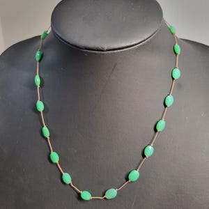 May include: A delicate necklace featuring oval-shaped, light green beads strung on a thin, gold-colored chain. The necklace is displayed on a black mannequin, showcasing the jewelry's design and color.