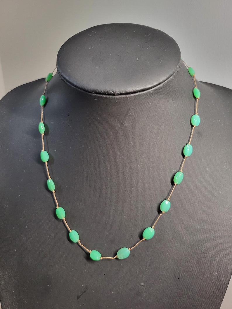 May include: A delicate necklace featuring oval-shaped, light green beads strung on a thin, gold-colored chain. The necklace is displayed on a black mannequin, showcasing the jewelry's design and color.