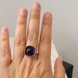 May include: A gold-colored ring featuring a large, square-cut, deep purple gemstone. The gemstone is surrounded by a decorative, textured gold setting. The ring is shown on a hand, with the focus on the jewelry.