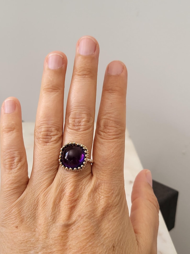 May include: A gold-colored ring featuring a large, square-cut, deep purple gemstone. The gemstone is surrounded by a decorative, textured gold setting. The ring is shown on a hand, with the focus on the jewelry.