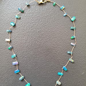 Op de afbeelding: Een delicate ketting met kleine, rechthoekige, iriserende opaal kralen in blauwe, groene en witte tinten, gelijkmatig verdeeld over een dun, lichtgekleurd koord. De ketting is voorzien van een goudkleurige metalen karabijnsluiting.