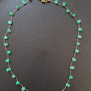 May include: A delicate necklace featuring small, teardrop-shaped green gemstones strung on a thin, gold-colored chain. The necklace has a gold clasp and is displayed against a dark background. The gemstones are evenly spaced along the chain.