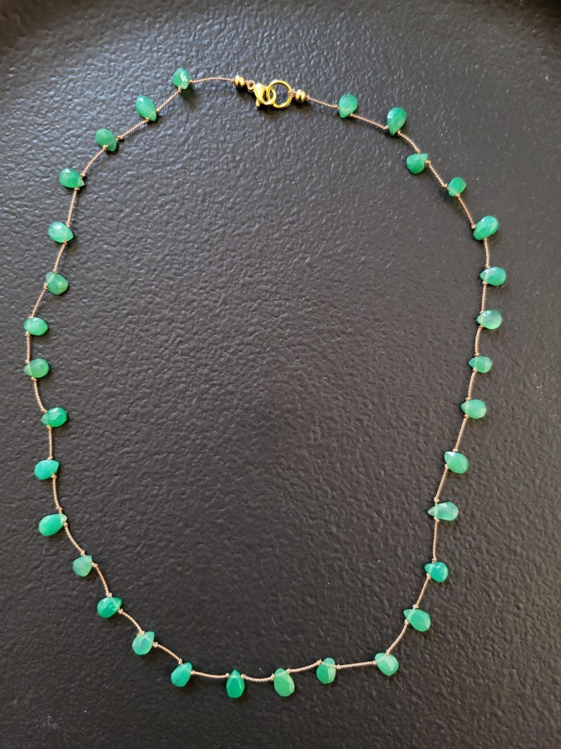 May include: A delicate necklace featuring small, teardrop-shaped green gemstones strung on a thin, gold-colored chain. The necklace has a gold clasp and is displayed against a dark background. The gemstones are evenly spaced along the chain.