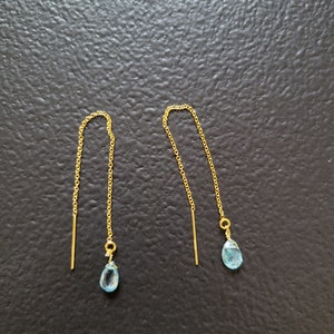 May include: A pair of gold-colored threader earrings. Each earring features a delicate gold chain with a small, light blue teardrop-shaped gemstone at the end. The earrings are displayed against a dark background.