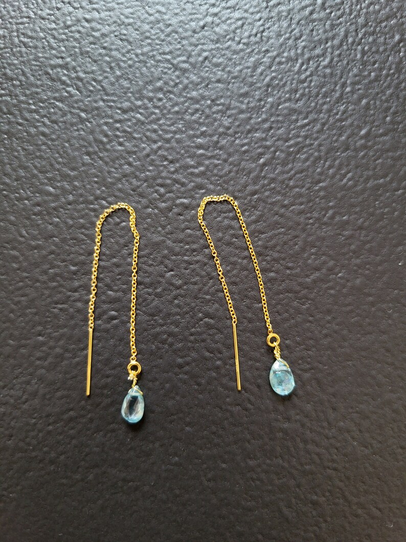 May include: A pair of gold-colored threader earrings. Each earring features a delicate gold chain with a small, light blue teardrop-shaped gemstone at the end. The earrings are displayed against a dark background.