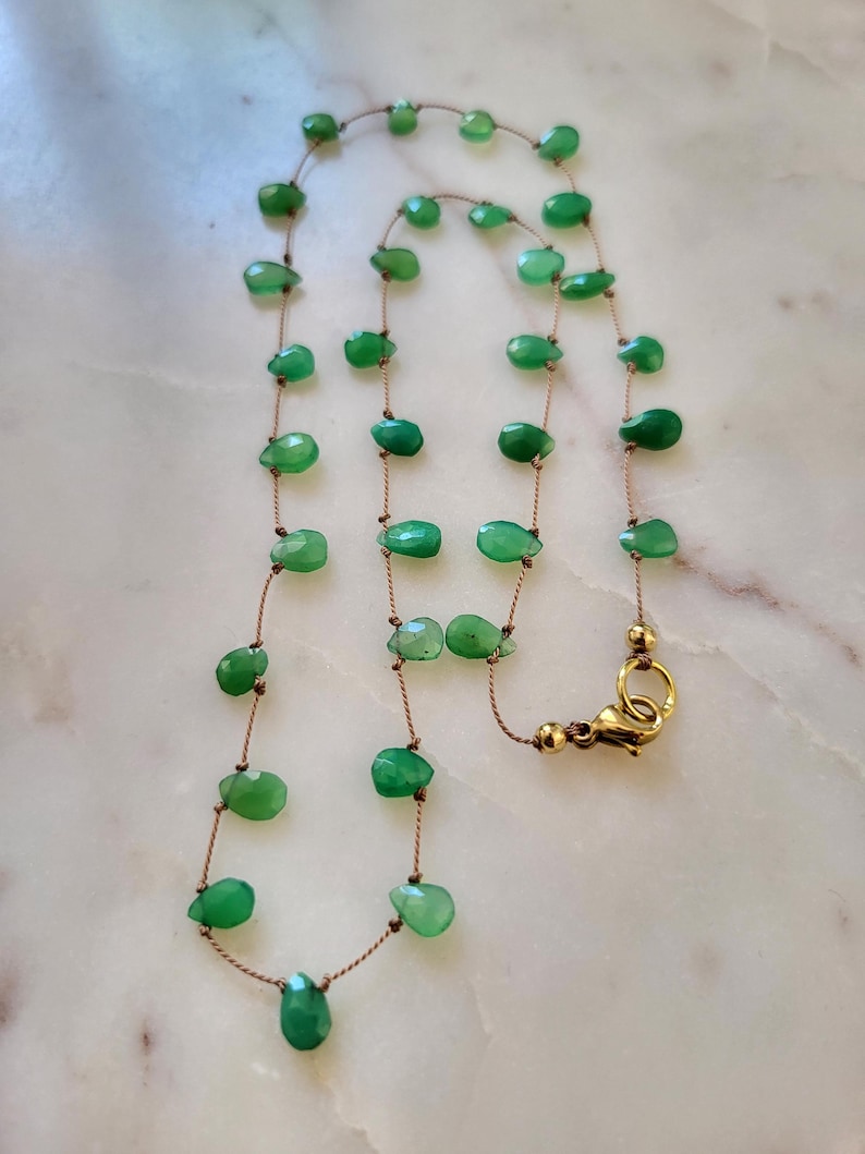May include: A delicate necklace featuring faceted, teardrop-shaped green gemstones strung on a thin, brown cord. The necklace has a gold-colored clasp. The gemstones are evenly spaced along the cord, creating a simple yet elegant design.