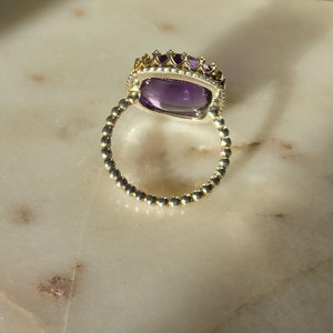May include: A gold-toned ring with a large, rectangular, purple gemstone. The ring features a beaded band and a crown-like setting around the gemstone. The ring is set on a light-colored, marble-like surface.