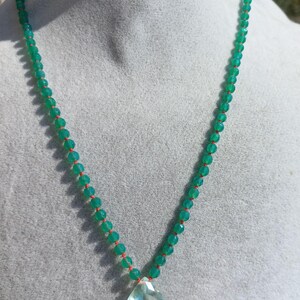 Può includere: Una collana composta da una fila di piccole perline verdi rotonde e sfaccettate infilate su un cordino arancione brillante. La collana include un unico grande ciondolo a forma di pera, sfaccettato e di colore verde chiaro, al centro.