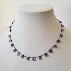 May include: A delicate necklace featuring small, teardrop-shaped, blue-gray beads strung along a dark cord. The beads are evenly spaced, creating a simple yet elegant design. The necklace is displayed on a white mannequin bust.