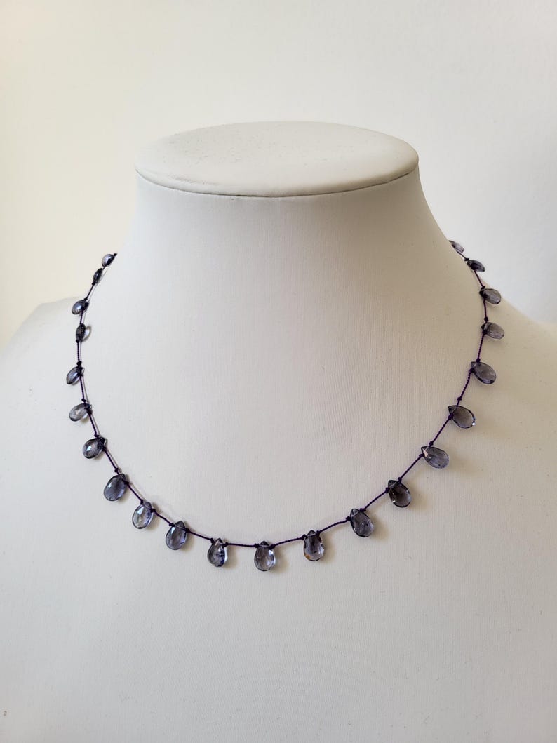 May include: A delicate necklace featuring small, teardrop-shaped, blue-gray beads strung along a dark cord. The beads are evenly spaced, creating a simple yet elegant design. The necklace is displayed on a white mannequin bust.