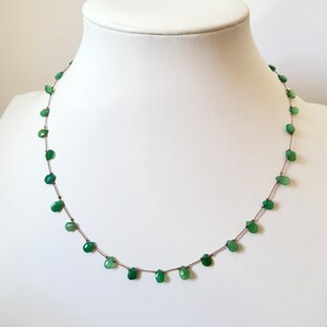 May include: A delicate necklace featuring small, teardrop-shaped green gemstones strung on a thin, brown cord. The gemstones are evenly spaced, creating a minimalist and elegant design. The necklace is displayed on a white mannequin bust.