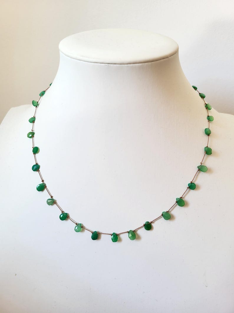 May include: A delicate necklace featuring small, teardrop-shaped green gemstones strung on a thin, brown cord. The gemstones are evenly spaced, creating a minimalist and elegant design. The necklace is displayed on a white mannequin bust.