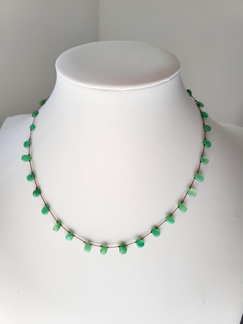 Pode incluir: Um delicado colar de pedras preciosas verdes com um cord&atilde;o marrom. O colar apresenta pequenas pedras preciosas de forma oval enfiadas em um cord&atilde;o fino.
