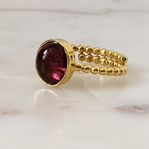 May include: A gold ring with a deep purple cabochon gemstone set in a bezel setting. The band is made of small gold beads.