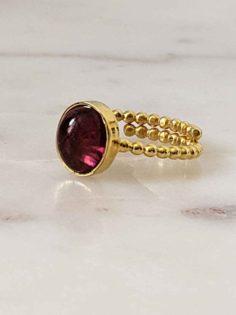 May include: A gold ring with a deep purple cabochon gemstone set in a bezel setting. The band is made of small gold beads.
