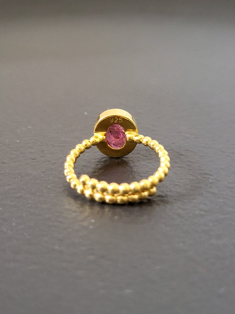 May include: A gold ring with a pink oval gemstone. The ring is made of a twisted band of small gold beads. The back of the ring is stamped with "925".