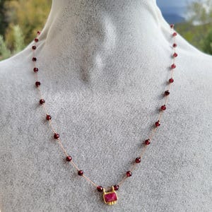 Puede incluir: Un delicado collar con pequeñas cuentas redondas de color rojo oscuro ensartadas en un fino alambre dorado. El collar tiene un colgante rectangular de color rubí con detalles dorados. El collar se exhibe en un maniquí gris.