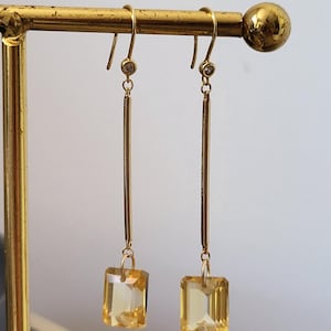 Peut inclure: Une paire de boucles d'oreilles pendantes en or composée d'une fine tige verticale suspendue à un petit clou circulaire orné d'une pierre claire. À la base de chaque tige pend une pierre jaune rectangulaire à facettes qui capte la lumière.