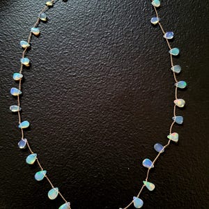 Può includere: Una collana delicata caratterizzata da una sottile catenina color oro con numerosi piccoli opali iridescenti a forma di goccia. Le pietre presentano una qualità traslucida con riflessi di luce blu e bianca, chiusa da un moschettone color oro.