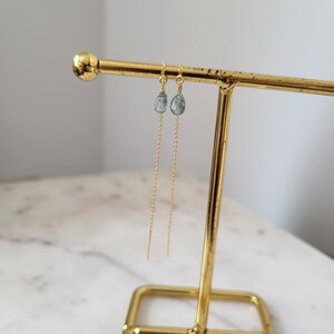 May include: A pair of gold-colored threader earrings with teardrop-shaped, pale blue gemstones. The earrings are displayed on a gold-colored jewelry stand against a white background. The gemstones appear to be approximately 0.25 inches in length.