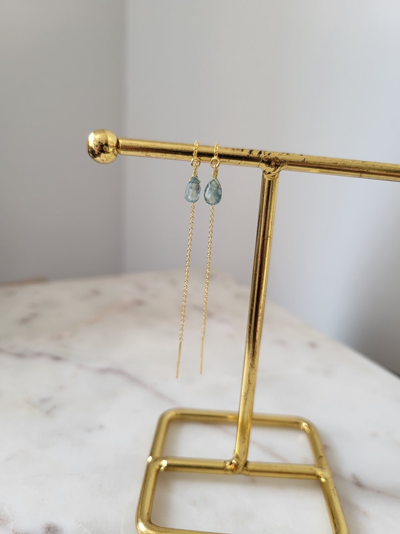 May include: A pair of gold-colored threader earrings with teardrop-shaped, pale blue gemstones. The earrings are displayed on a gold-colored jewelry stand against a white background. The gemstones appear to be approximately 0.25 inches in length.