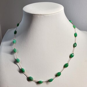 May include: A delicate necklace featuring oval-shaped, bright green beads strung on a thin, brown cord. The beads are evenly spaced, creating a minimalist and elegant design. The necklace is displayed on a white mannequin bust.