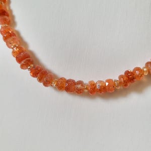 Peut inclure: Un collier compos&eacute; de perles de pierre de soleil orange-rouge &agrave; facettes. Les perles sont enfil&eacute;es, cr&eacute;ant un bijou d&eacute;licat et scintillant. Le collier est pr&eacute;sent&eacute; sur un fond blanc uni.