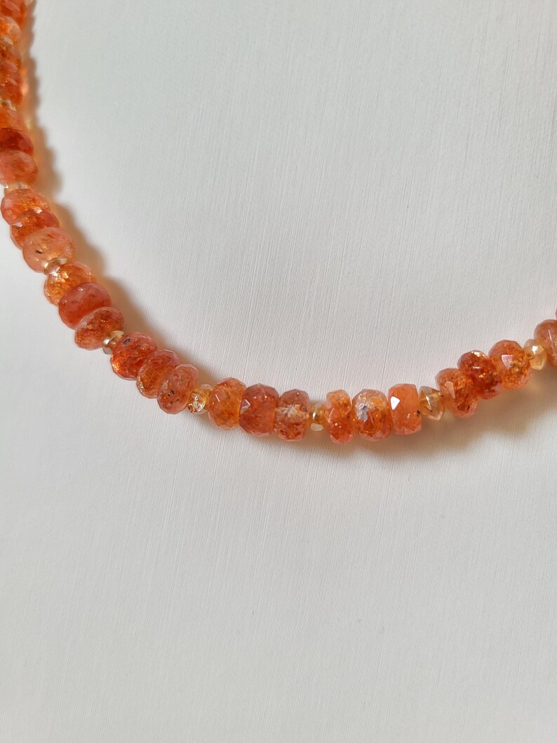 Peut inclure: Un collier compos&eacute; de perles de pierre de soleil orange-rouge &agrave; facettes. Les perles sont enfil&eacute;es, cr&eacute;ant un bijou d&eacute;licat et scintillant. Le collier est pr&eacute;sent&eacute; sur un fond blanc uni.
