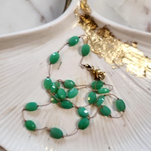 May include: A delicate necklace featuring faceted, oval-shaped green beads strung on a thin, gold-colored chain. The necklace is displayed on a white, shell-shaped dish with gold leaf accents. The clasp is gold-toned.